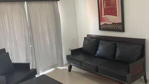 Una sala de estar moderna con un sofá negro, una mesita de centro y una silla. Hay un ventanal con cortinas translúcidas y una obra de arte enmarcada en la pared.