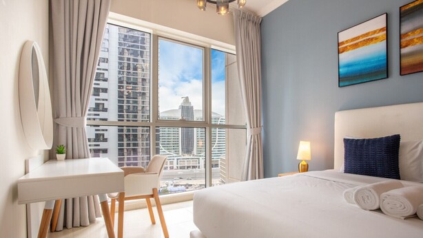 A hotel room with a large bed, a desk with a chair, a mirror, and a view of a cityscape through large windows. - Premium 1 Bed | Walk to Dubai Mall, Burj Khalifa (Downtown Dubai)