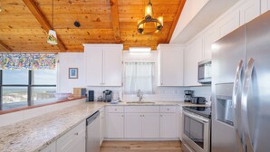 A kitchen with white cabinets, stainless steel appliances, a wooden ceiling, and a view through the window. - Conch Out 1405 | Gulf-Front Beach House (Gulf Shores)