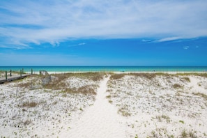 Una playa con un paseo marítimo de madera que conduce al agua, dunas arenosas y un cielo azul despejado.