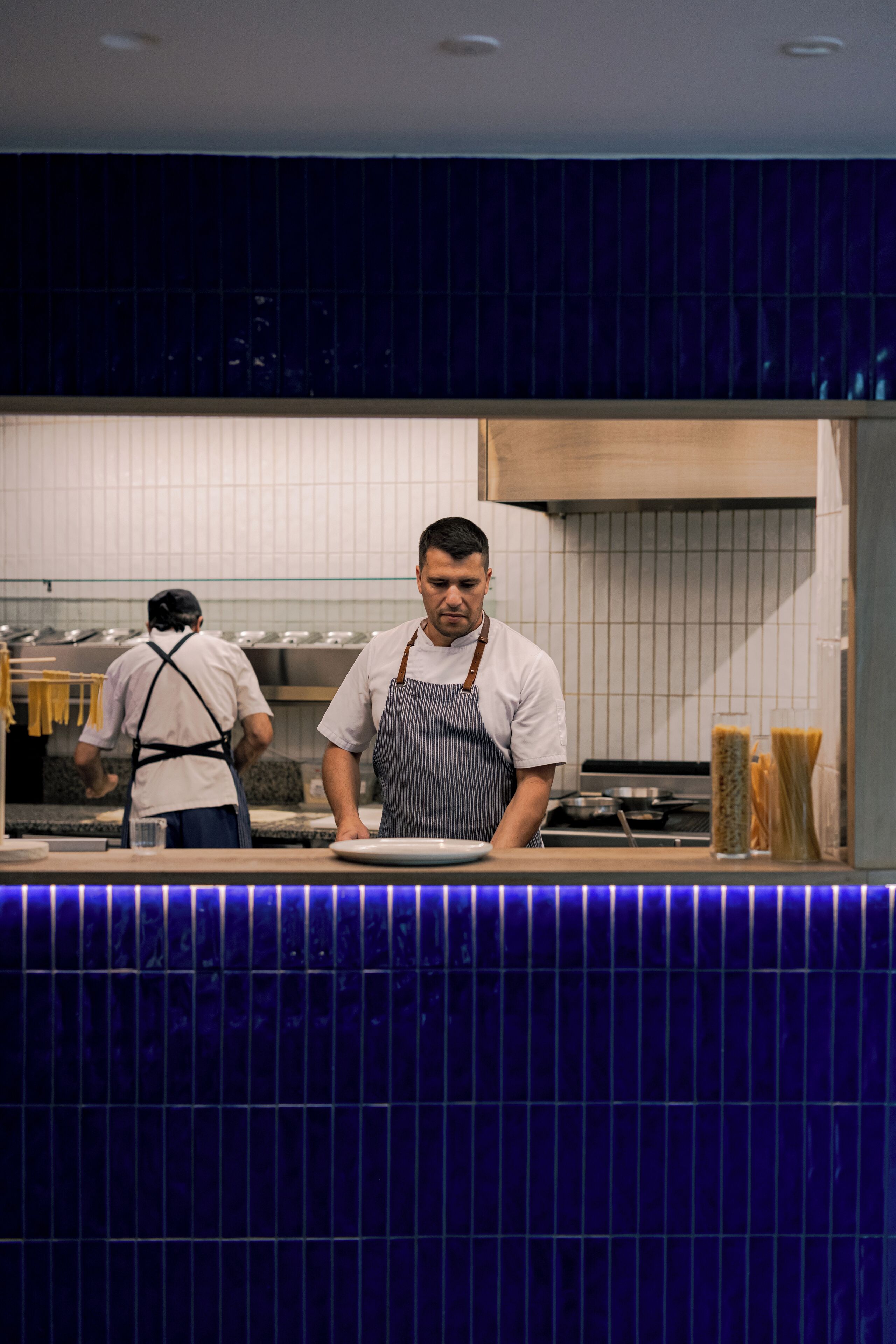 A kitchen with two chefs, one in the foreground and one in the background, with a tiled backsplash and stainless steel appliances.