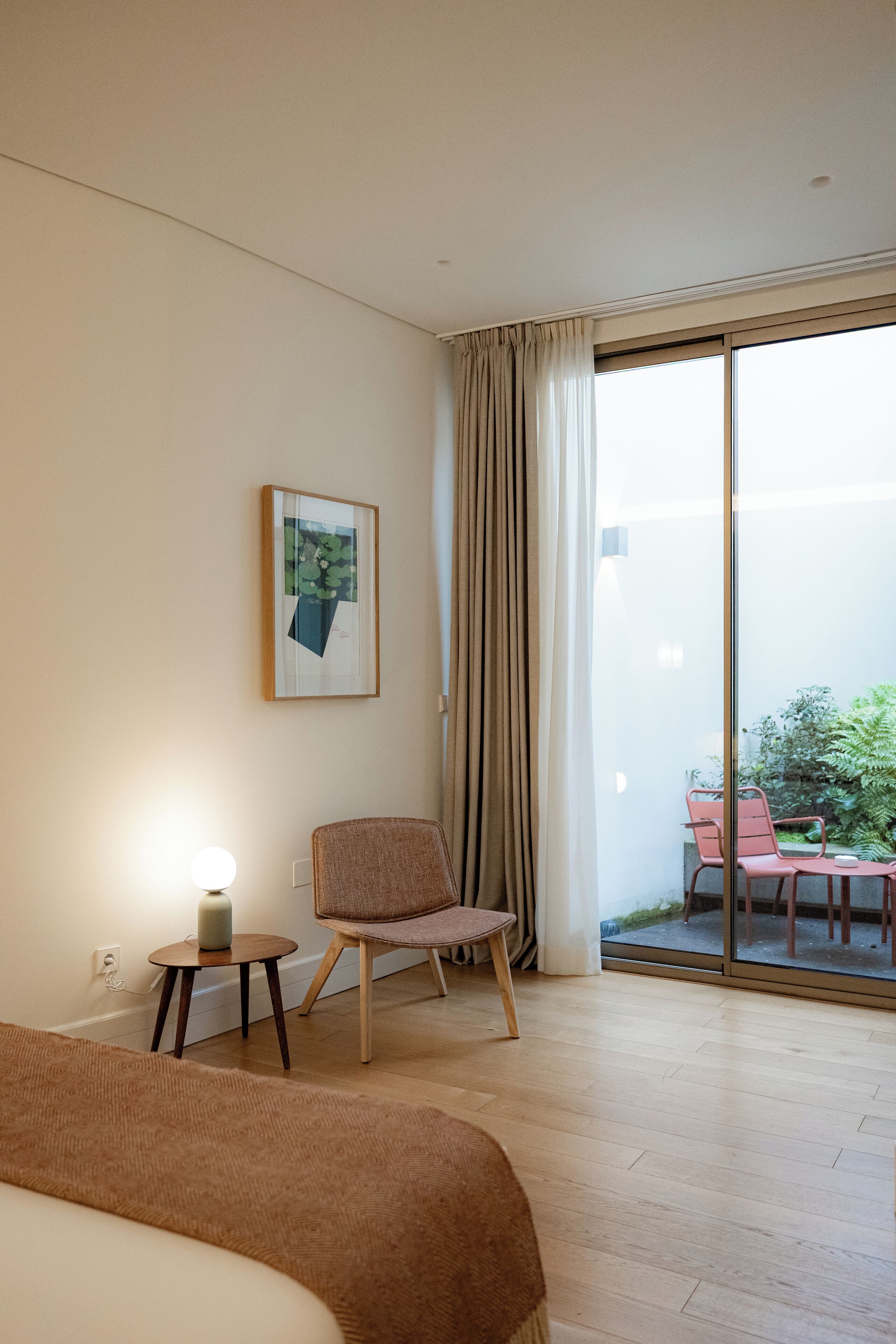 Habitación de hotel con cama, una silla, una mesita de noche con lámpara, un cuadro en la pared y puerta corrediza de vidrio que da a un espacio exterior con mesa y sillas.
