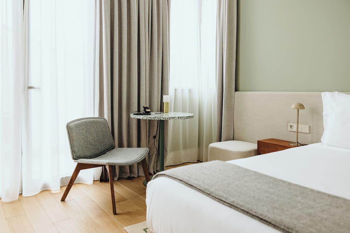 A hotel room with a bed, a chair, a small table with a lamp, and a nightstand.