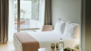 A hotel room with a large bed, a bedside table with a bottle, glasses, and a bowl of fruit, and a view of the outdoors through a sliding door.