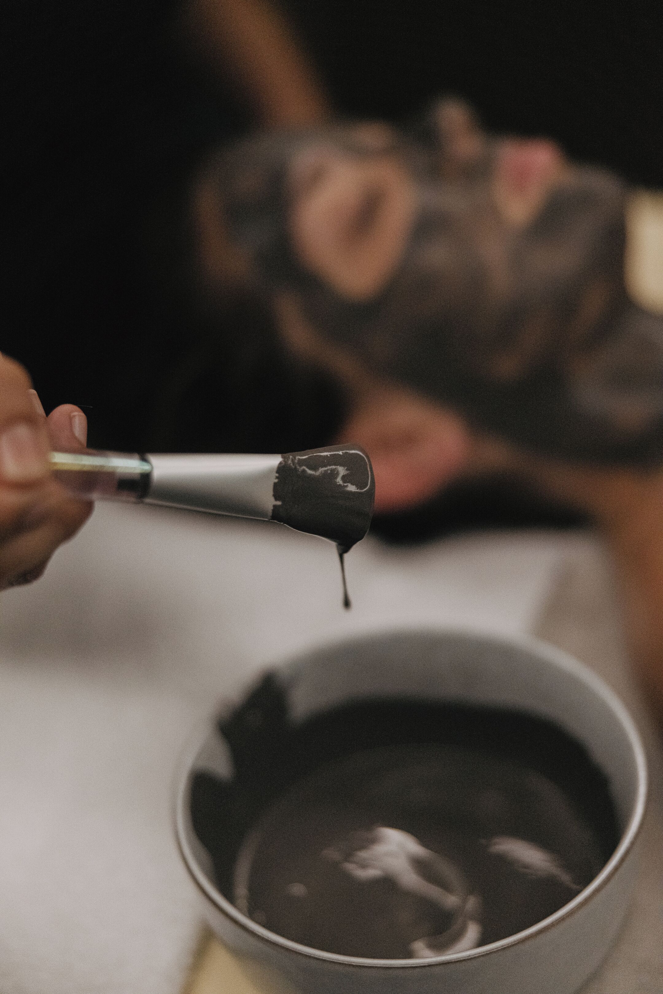 A close-up of a person holding a brush with paint dripping from it, with a blurred background of another person.