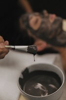 A close-up of a person holding a brush with paint dripping from it, with a blurred background of another person.