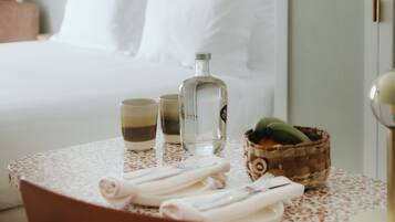 Una cama bien hecha con sábanas blancas, una mesita con mantel de encaje, una botella, dos vasos, un cuenco y una cesta con frutas.