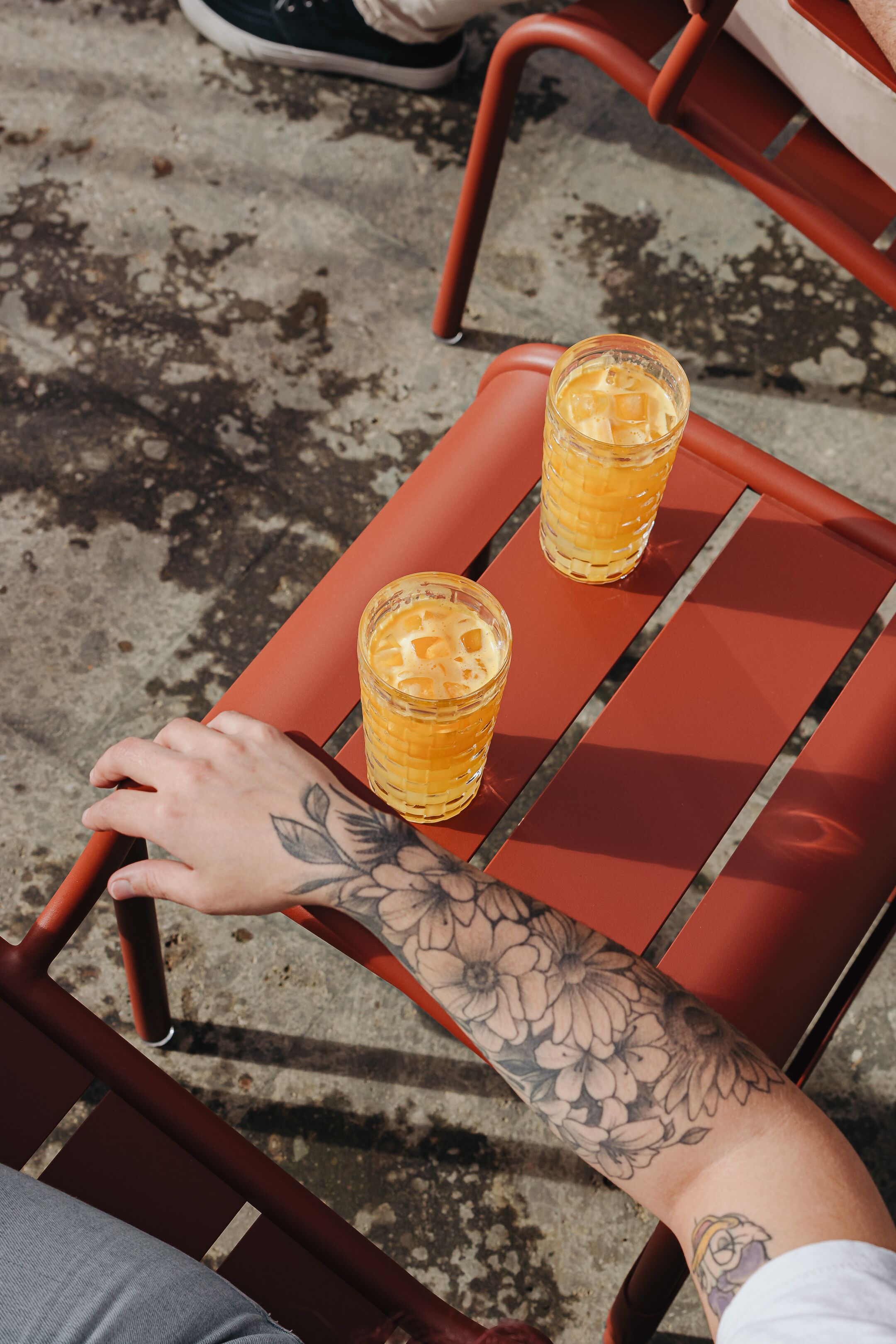 En person med en tatuering på armen sitter på en röd stol, och på ett bord framför personen står två glas med en guldryck.
