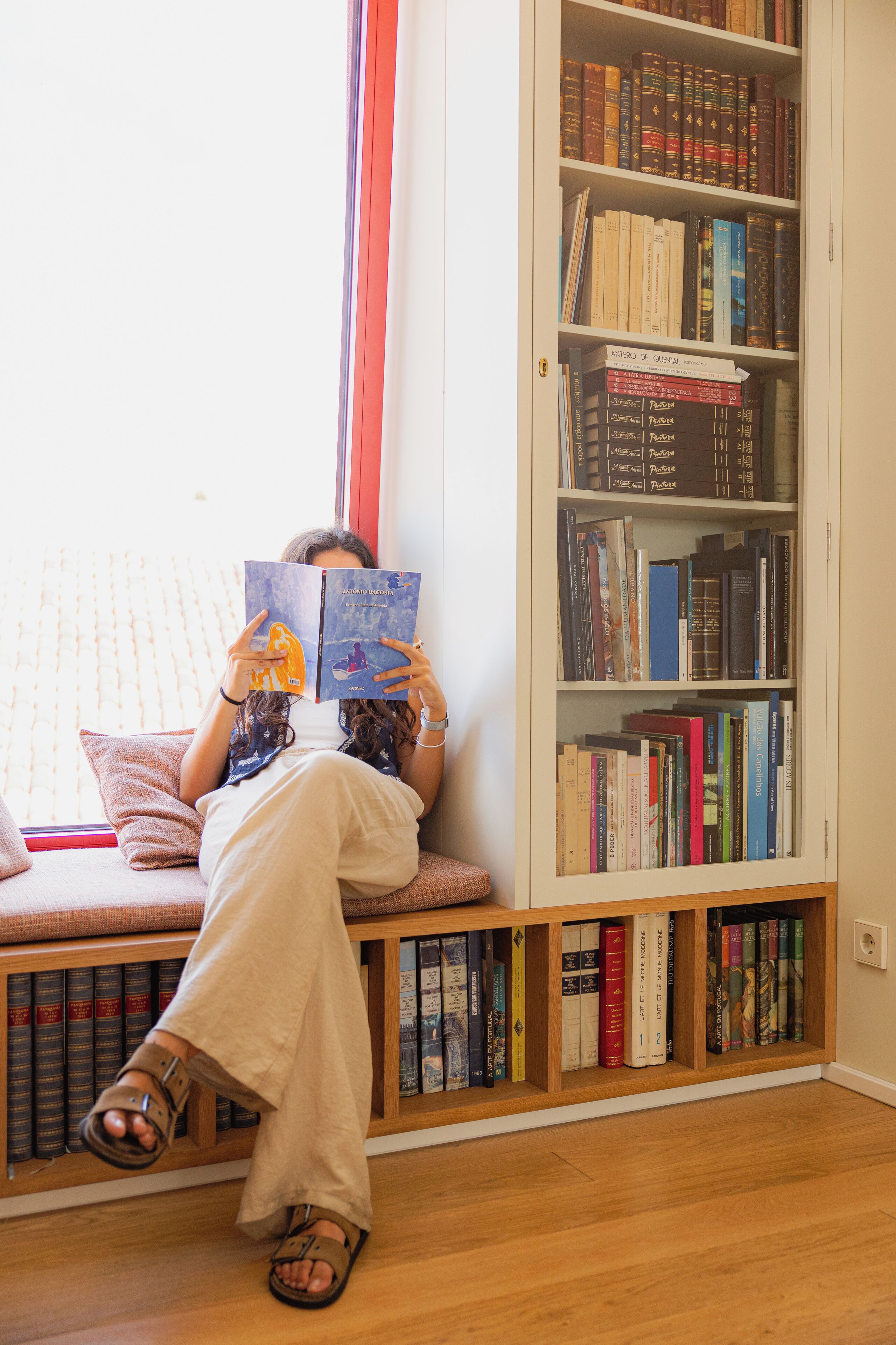 A person reading a book on a bench in a room with a bookshelf.