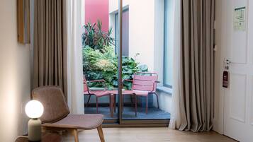 Una habitación con piso de madera, una silla, una mesa y un ventanal corredero que da a un espacio exterior con plantas y una pared roja.