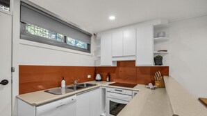 A compact kitchen with white cabinets, a sink, a stove, and a countertop with a kettle and a bottle. - Warrina 8 | Thredbo | Large & on the Village flat (Thredbo)