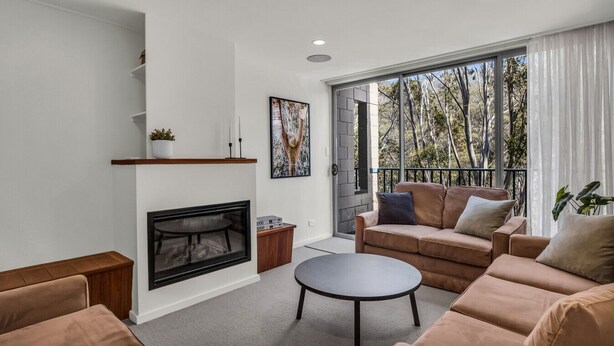 A modern living room with a fireplace, a sofa set, a coffee table, and a large window with a view of trees. - Warrina 8 | Thredbo | Large & on the Village flat (Thredbo)