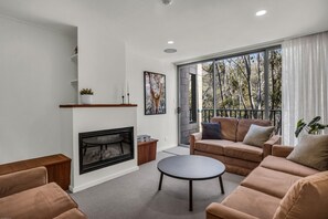 A modern living room with a fireplace, a sofa set, a coffee table, and a large window with a view of trees. - Warrina 8 | Thredbo | Large & on the Village flat (Thredbo)