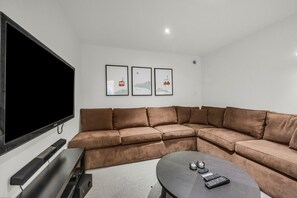 Una sala de estar moderna con un televisor de pantalla plana grande, un sofá seccional marrón, una pequeña mesa redonda y tres cuadros en la pared.
