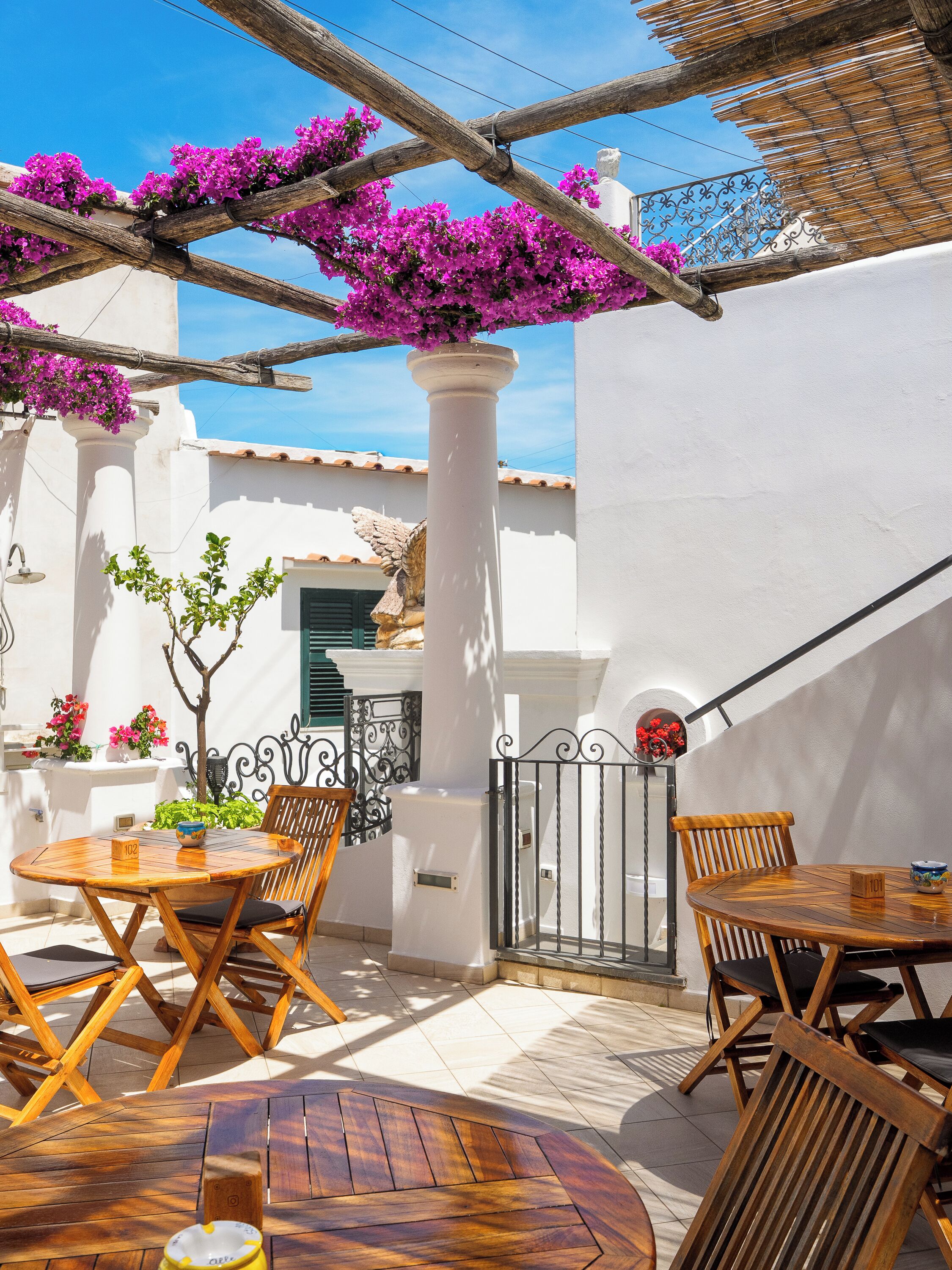An outdoor patio with wooden tables and chairs, a white column, and purple bougainvillea flowers.