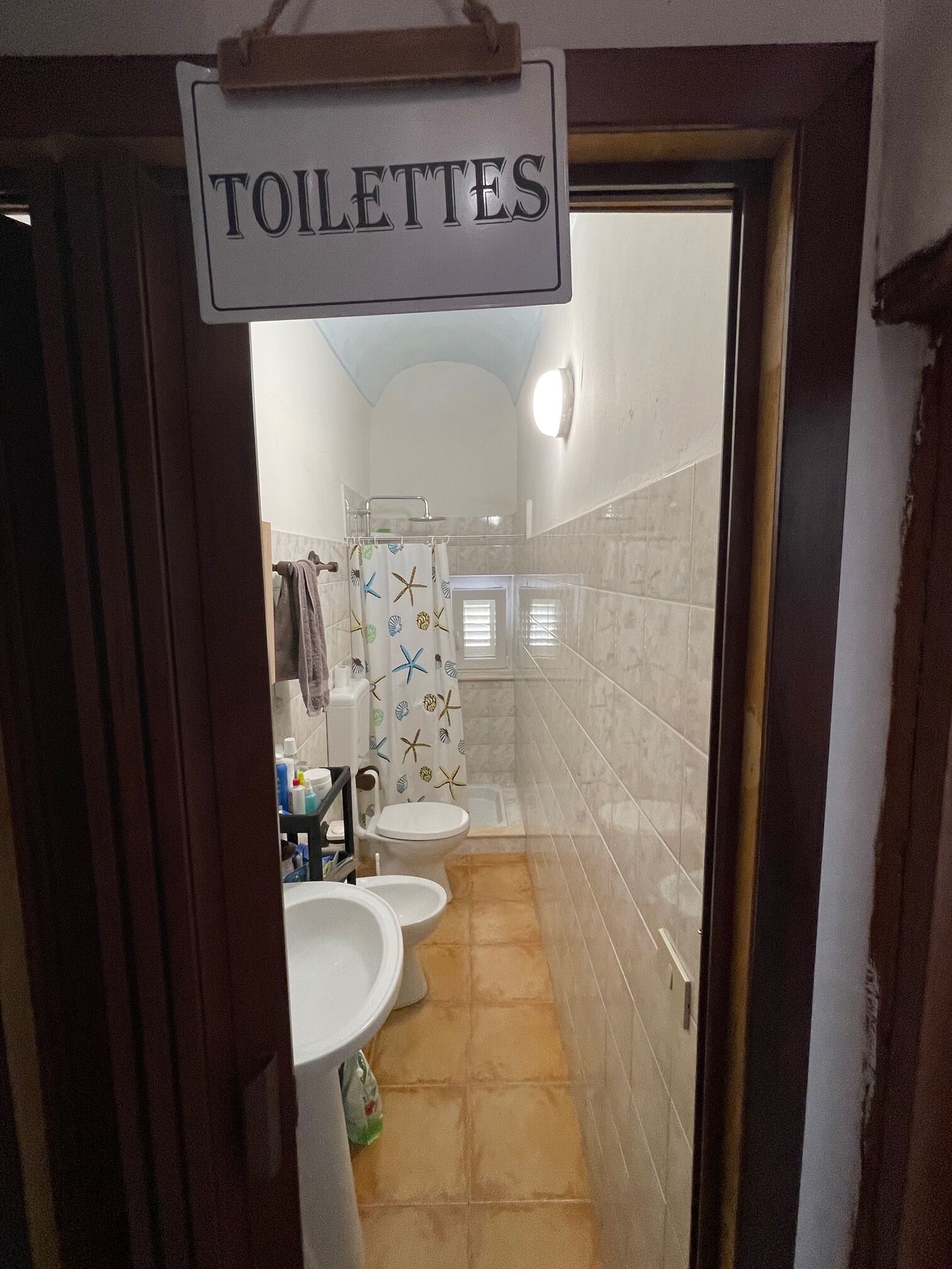 Um banheiro com duas pias, vaso sanitário e chuveiro. Há uma placa acima da porta com a inscrição 'TOILETTES'.