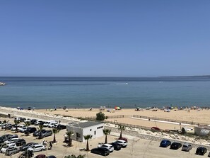 A beachside parking lot with cars and a small building, overlooking a sandy beach with umbrellas and people.