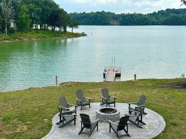 Une terrasse au bord du lac, avec des fauteuils Adirondack et un foyer, offrant une vue sur un lac paisible et un quai.