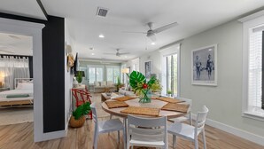 A modern living room with a dining area, featuring a round table with white chairs, a large plant centerpiece, and a ceiling fan.