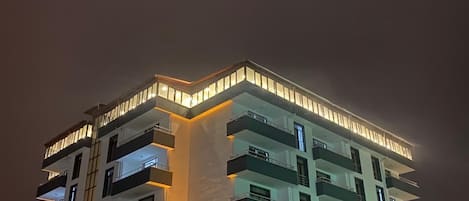 Un edificio de varios pisos con balcones, ventanas iluminadas y suelo cubierto de nieve.