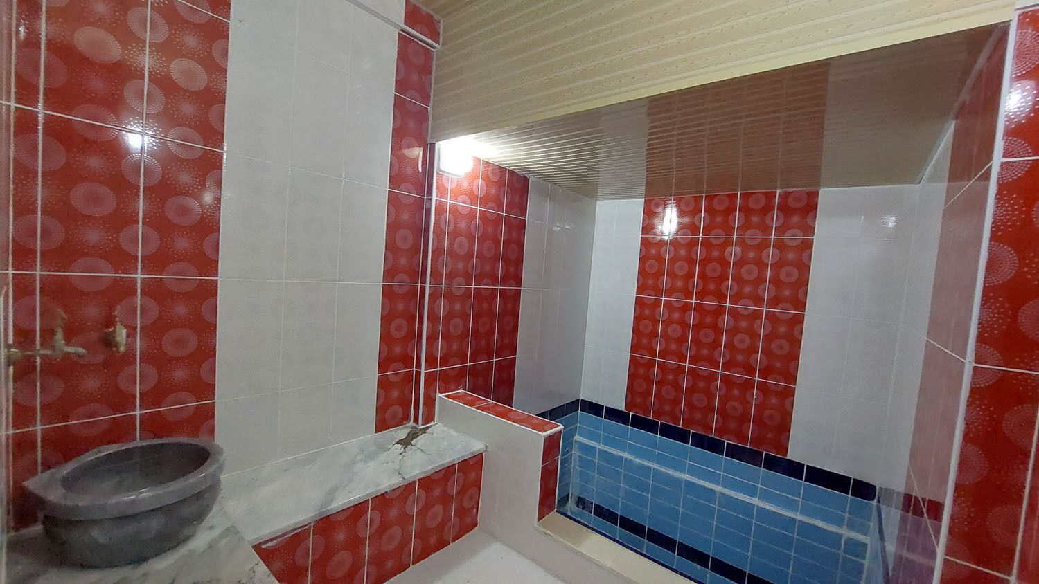 Une salle de bain avec des carreaux rouges et blancs formant un motif, un lavabo en marbre et une zone de douche carrelée de bleu.