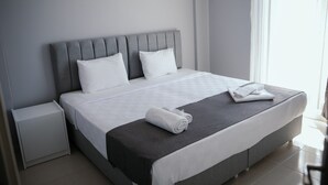 Una cama bien hecha con sábanas blancas y un cabecero gris, una mesita de noche pequeña y una ventana con cortinas de encaje.
