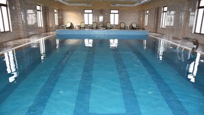 Una gran piscina interior con agua cristalina azul, paredes con azulejos y varias sillas de descanso a lo largo de los bordes.