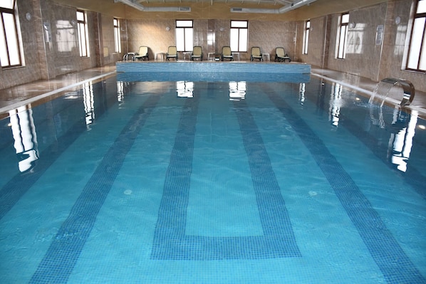 Une grande piscine intérieure à l’eau d’un bleu limpide, avec des murs carrelés et plusieurs chaises longues disposées le long des bords.