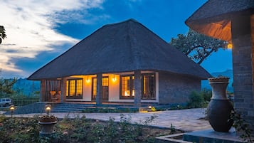 A thatched-roof building with a stone facade, illuminated windows, and a paved entrance area with a large vase.