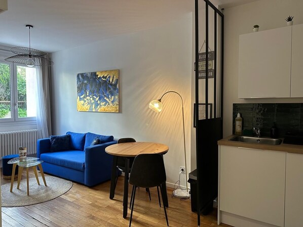 A modern living room with a blue sofa, a round wooden table, a floor lamp, and a kitchen area with a sink and cabinets.