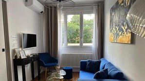 Ein modernes Wohnzimmer mit einem blauen Sofa, einem kleinen Tisch, einem Sessel, einem Fernseher und einem Fenster mit Blick auf Grünflächen.