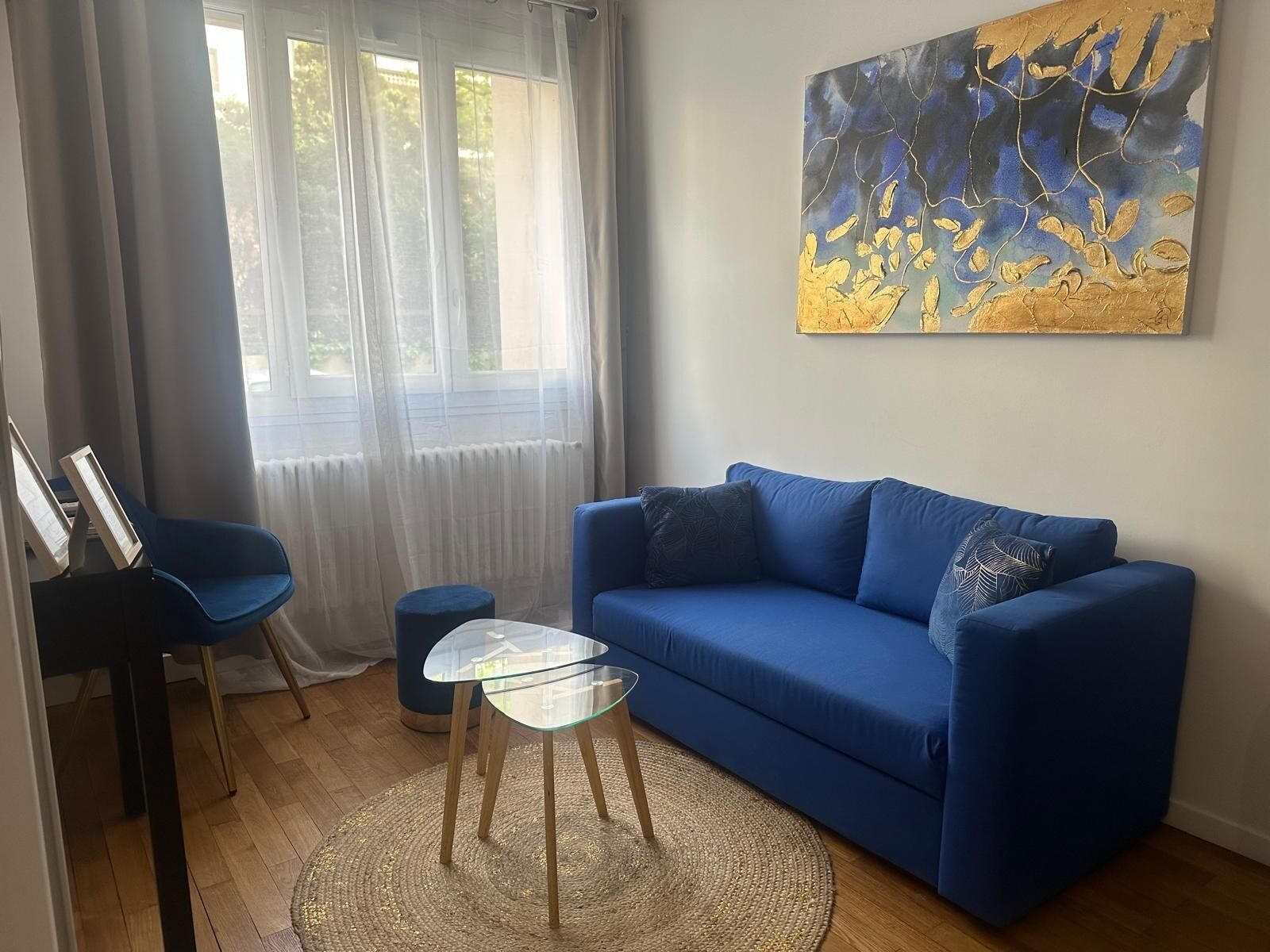 A living room with a blue sofa, a glass coffee table, a blue chair, and a painting on the wall.