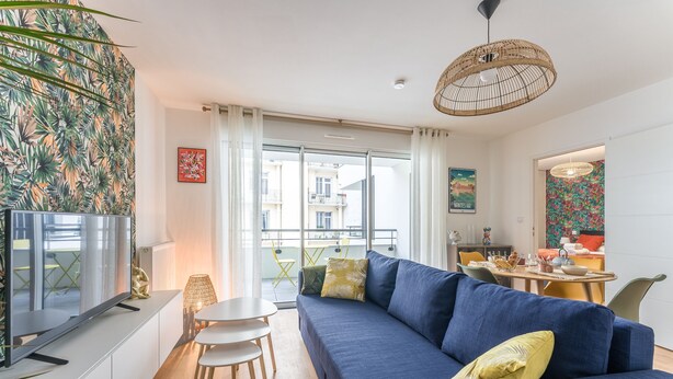 Una sala de estar moderna con un sofá azul, un televisor de pantalla plana, un comedor con mesa y sillas, y un balcón con vistas a edificios.