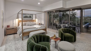 A modern bedroom with a four-poster bed, a wooden nightstand, a green armchair, and a glass door leading to a balcony with a view of the outdoors.
