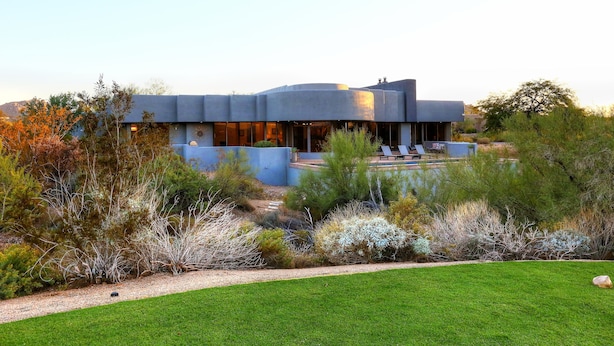 A modern building with a curved roof and large windows, surrounded by a well-maintained garden and a small pool area.