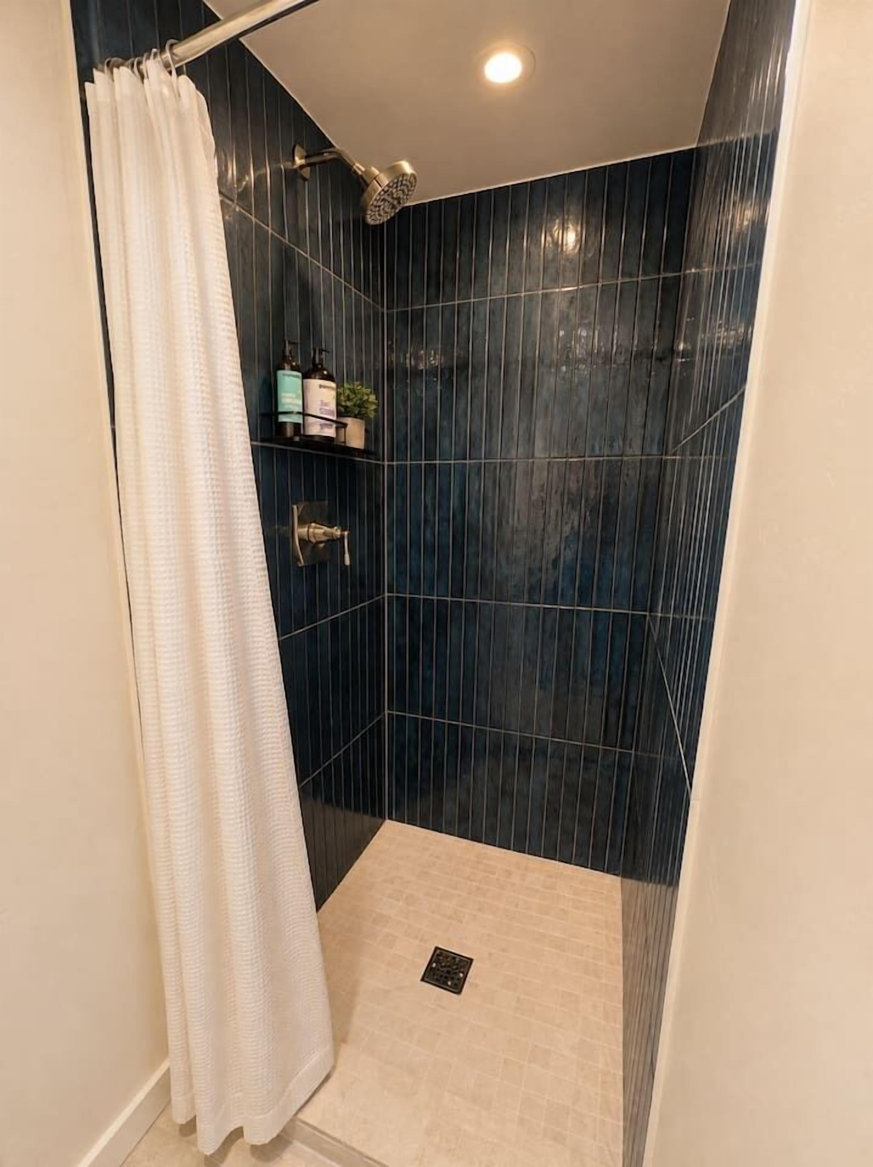 A shower area with dark blue tiles, a white shower curtain, and a small shelf with toiletries.
