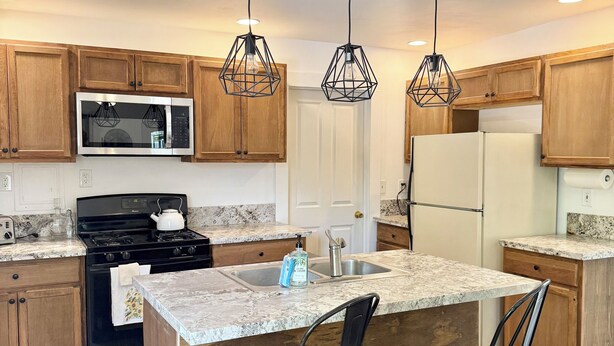 A modern kitchen with wooden cabinets, a stainless steel microwave, a black stove, and a white refrigerator. There are two bar stools and pendant lights above the island. - Spacious 3 Bedroom Home with yard, pet friendly & sleeps 6! (Oroville)