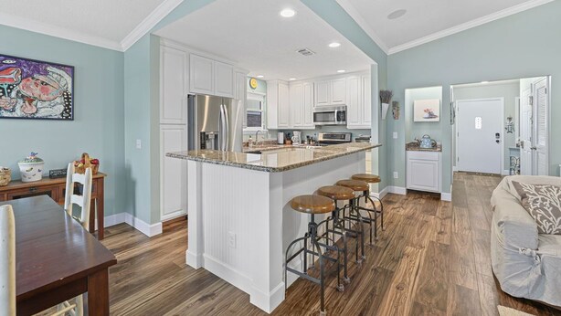 A modern kitchen with white cabinets, a central island with stools, and a dining area with a wooden table and chairs. - Pool Home Walk to Beach | Good Morning Rentals (Jacksonville Beach)