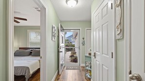 A hallway with a wooden floor, a bed with gray bedding, and a door leading outside. - Pool Home Walk to Beach | Good Morning Rentals (Jacksonville Beach)