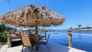 A wooden deck with a thatched umbrella, a table, and chairs overlooking a calm body of water. - Prestige Waterfront Paradise: Sunset Views, Private Pool & Boat Rental Option! (Cape Coral)