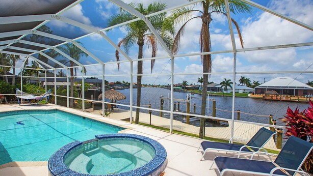 A pool area with a hot tub, lounge chairs, and a view of a body of water with palm trees. - Prestige Waterfront Paradise: Sunset Views, Private Pool & Boat Rental Option! (Cape Coral)