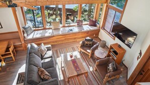 Ein gemütliches Wohnzimmer mit Holzbalkendecke, großen Fenstern, einer Couch, einem Couchtisch und einem Fernseher.