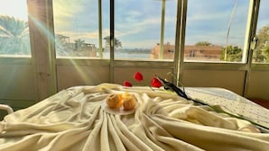 Una habitación con una cama cubierta con una manta blanca, una ventana con vista a un paisaje urbano y un jarrón con flores rojas y un par de pantuflas sobre la cama.