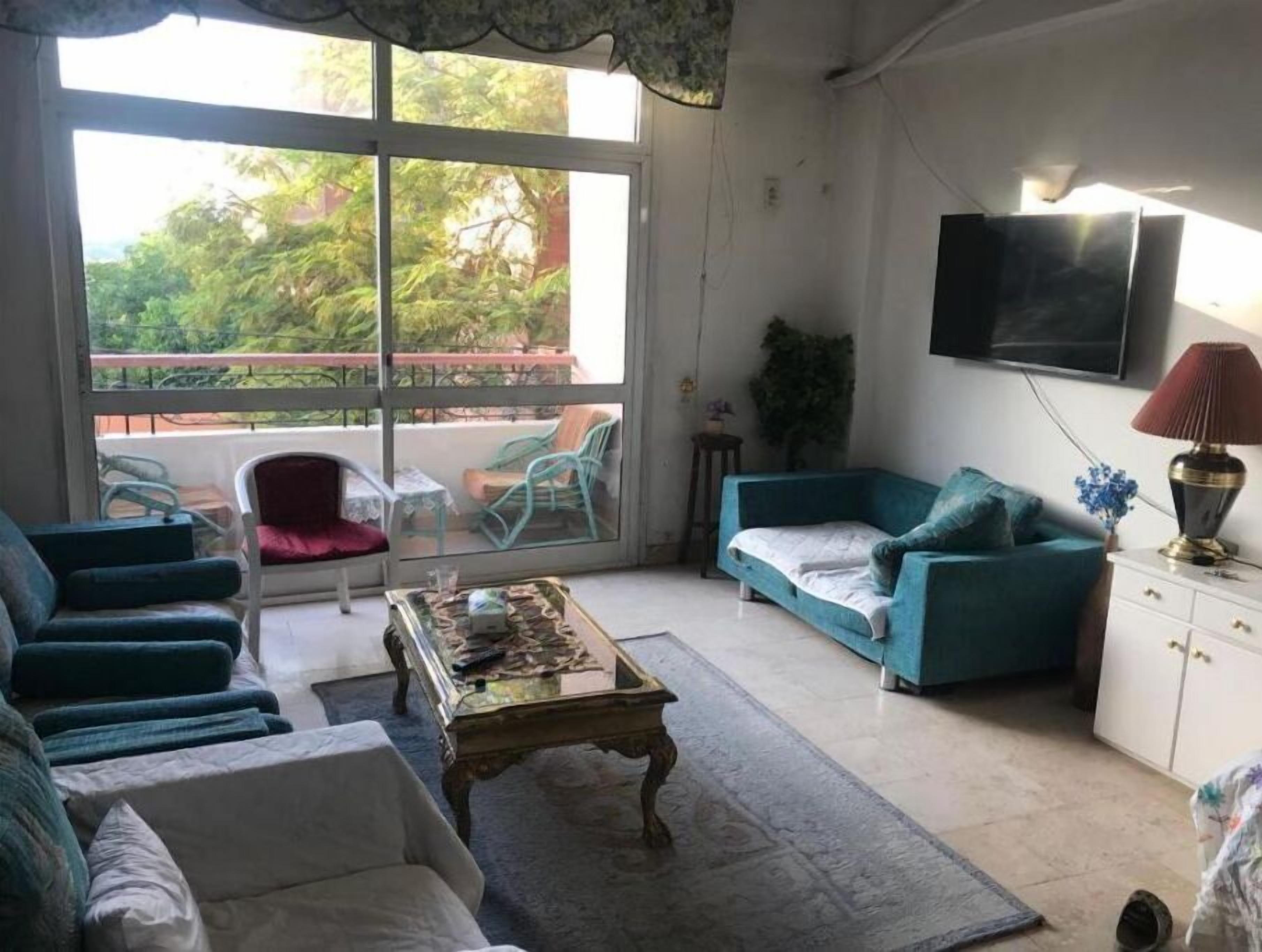 Un salón con sofá, una mesa de centro y televisor en la pared. Tiene un balcón con sillas y vistas a zonas verdes.