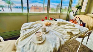 A room with a table set for breakfast, a view of palm trees and buildings, and a window with curtains. - Luxor city A block in front of NILE RIVER 3 (Luxor City)