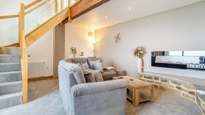 A modern living room with a grey sofa, wooden coffee table, and a stone accent wall. A wooden staircase with glass railings leads to an upper level. - 2 bedroom accommodation in Carleton, near Skipton (Carleton, near Skipton)