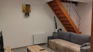 Ein Wohnzimmer mit grauem Sofa, einem Holztisch, einem Fernseher auf einem Holzboden, einem Heizkörper, einer Treppe und einem Gemälde an der Wand.