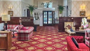 A spacious lobby with a red patterned carpet, a red sofa, a beige armchair, and a red velvet sofa. There are potted plants and wall-mounted lamps.