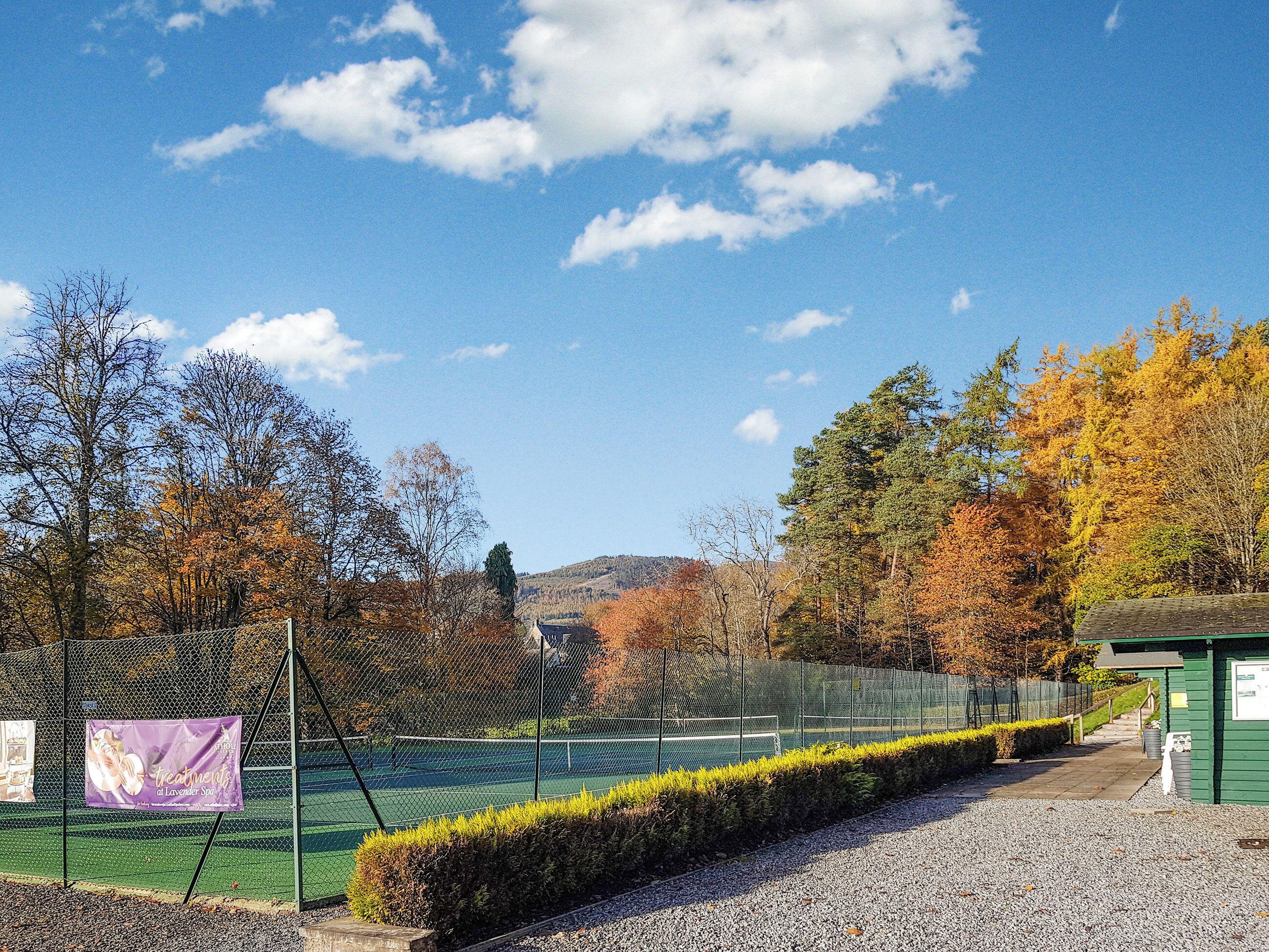 A tennis court with a fence, a small pavilion, and autumn trees in the background.