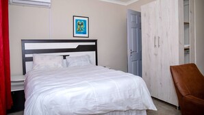 Una cama bien tendida con ropa blanca, cabecera de madera, cortina roja y una silla de cuero marrón.